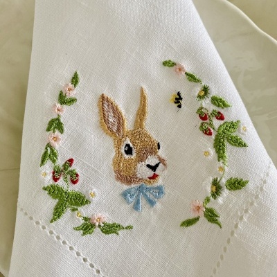 Guardanapo branco com bordado de coelho, flores e frutos vermelhos sobre prato creme
