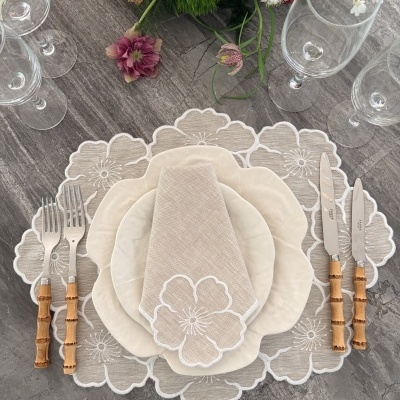 Mesa posta com pratos creme, talheres com cabos de bambu e guardanapo bege bordado com flores.