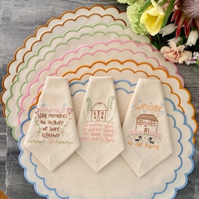Guardanapos de tecido bordados com frases e flores sobre toalhas redondas coloridas numa mesa de pedra cinzenta ao lado de flores.