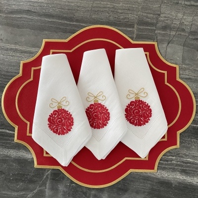 Guardanapos brancos bordados com bolas de Natal vermelhas, sobre jogo americano vermelho com borda dourada.