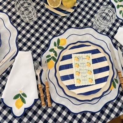 Mesa com toalha xadrez azul e branca, pratos risca azul e creme, guardanapo com bordado limão, talheres bambu, copos transparentes com padrão branco
