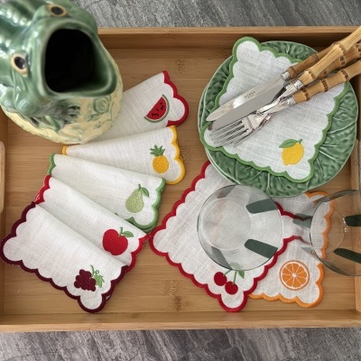 Tabuleiro com jarra cerâmica, pratos verdes, guardanapos com bordados de frutas, talheres com cabos de bambu e copos transparentes