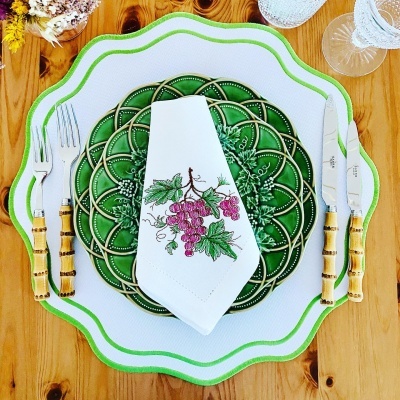 Mesa posta com pratos verdes e brancos, talheres com cabo de bambu e guardanapo bordado com uvas
