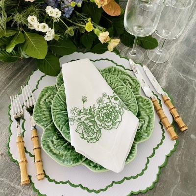 Mesa posta com pratos verdes, guardanapo branco bordado, talheres com cabo de bambu, copos de vidro e flores