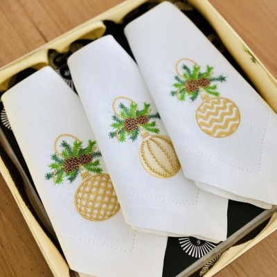 Três guardanapos brancos com bordados de bolas de Natal douradas e verdes numa caixa amarela