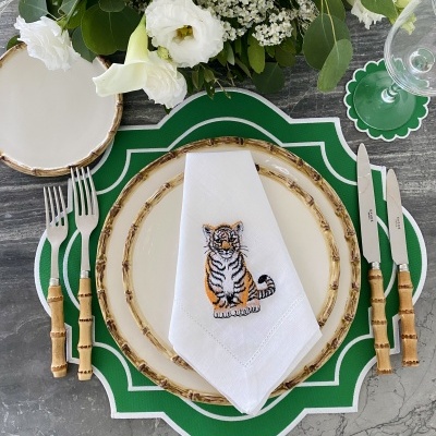 Mesa posta com pratos com borda de bambu, talheres com cabos de bambu, guardanapo branco bordado com tigre, base de prato verde e flores brancas no centro
