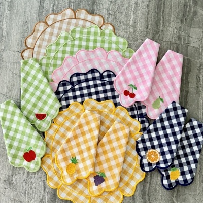 Guardanapos e bases de mesa xadrez com bordas onduladas e detalhes de frutas bordadas em tecido sobre superfície cinza.