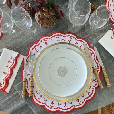 Mesa com serviço de jantar branco e dourado, base decorativa vermelha, talheres com cabo de bambu, copos transparentes e decoração floral seca