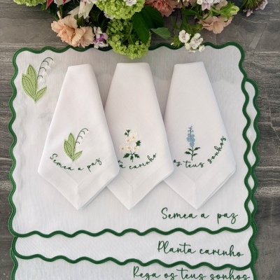 Jogo de toalhas de pano brancas com bordados de flores e texto verde, sobre toalha maior com bordado e acabamento ondulado verde.