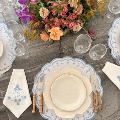 Mesa posta com louça branca em formato floral, talheres com cabo de bambu, guardanapos bordados e arranjo de flores coloridas