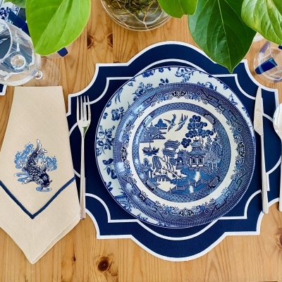 Prato de porcelana azul e branco com padrão oriental, guardanapo bege com bordado azul, talheres e copos sobre mesa de madeira com folhas verdes