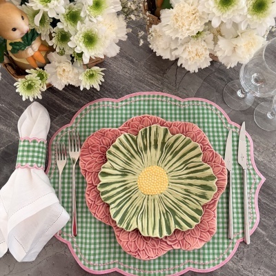 Mesa posta com pratos em forma de flor, toalhete xadrez verde e branco, talheres, guardanapo branco e flores brancas.