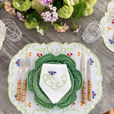 Mesa posta com pratos verdes em forma de folhas, guardanapo branco bordado e talheres com cabo amarelo sobre toalha branca bordada e arranjo floral colorido.