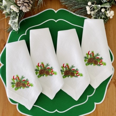 Quatro guardanapos brancos bordados com pinhas e fitas vermelhas sobre placemat verde com decoração de inverno
