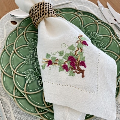 Guardanapo branco bordado com ramo de uvas sobre prato verde cerâmico, guardanapo preso por argola de madeira trançada numa mesa de madeira clara