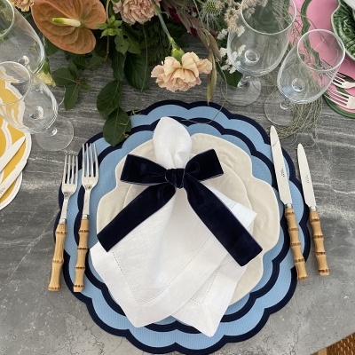 Mesa posta com base azul, prato branco, guardanapo branco com laço azul, talheres com cabo bambu e arranjo floral