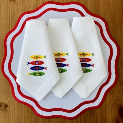 Guardanapos brancos com bordado colorido de peixes sobre tabuleiro branco com borda vermelha