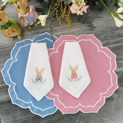 Duas peças de tecido branco com bordado de coelhos, sobre suportes de mesa rosa e azul, com decoração primaveril