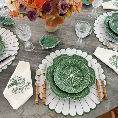 Mesa posta com pratos verdes de formato folha, guardanapos bordados com pássaros verdes, copos de vidro, talheres com cabos de madeira e centro de flores coloridas.