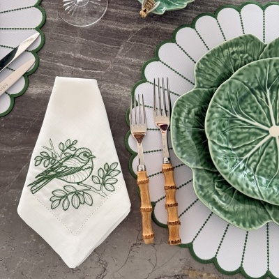 Mesa de jantar com loiça verde em forma de folha, talheres com cabo de bambu, e guardanapo branco bordado com pássaro verde