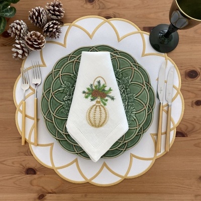 Mesa posta com prato verde, guardanapo branco bordado, talheres dourados, placemat branco e dourado, copo verde e pinhas decorativas.