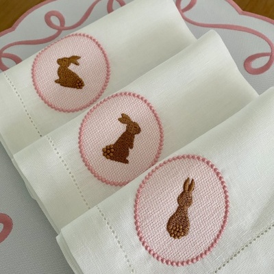 Guardanapos brancos com bordados de coelhos castanhos em oval cor de rosa sobre superfície branca com padrão rosa