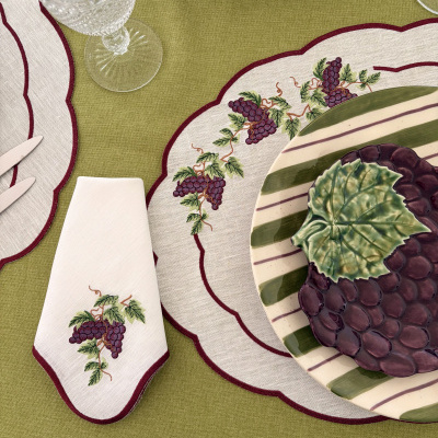 Conjunto de mesa com pratos decorados, guardanapo e jogo americano bordados, e copo de vidro texturizado sobre toalha verde.
