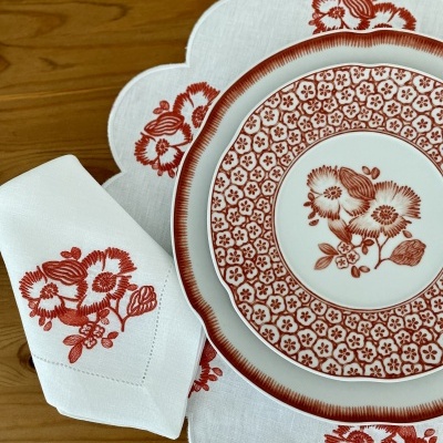 Conjunto de jantar com prato, guardanapo e toalha branca com padrões florais vermelhos sobre mesa de madeira