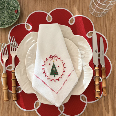 Conjunto de mesa com pratos brancos texturizados, guardanapo bordado de Natal, talheres com cabo em bambu, base de tecido vermelha e copo de vidro.