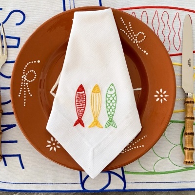 Conjunto de mesa com prato castanho decorado, guardanapo branco bordado com peixes multicoloridos, faca com cabo de madeira e toalha bordada colorida