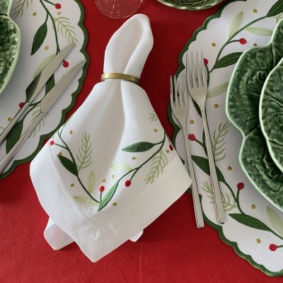 Mesa posta com pratos e guardanapo decorados com padrão de folhas e bagas em verde e vermelho, sobre toalha vermelha.