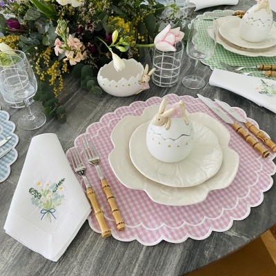 Mesa com jogo americano rosa xadrez, prato em forma de flor, ovo decorativo com coelho, talheres de cabo madeira e jarro de flores.