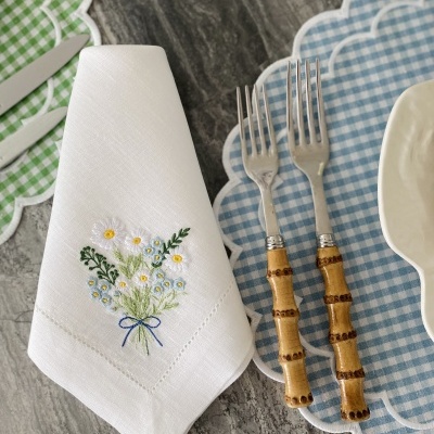 Guardanapo branco bordado com flores ao lado de garfos com cabo em bambu e prato sobre sousplat azul xadrez