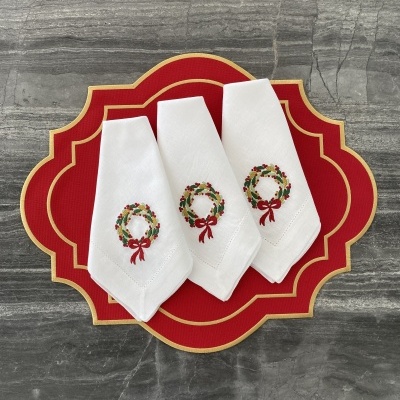 Guardanapos brancos com coroa de Natal bordada sobre descanso de prato vermelho com borda dourada
