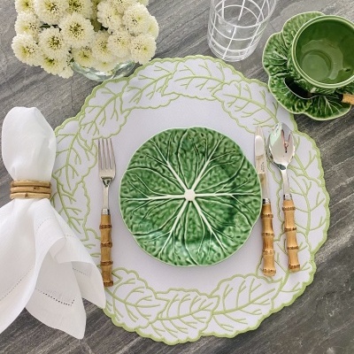 Mesa posta com prato verde de folha, prato branco com folhas desenhadas, talheres com cabo de bambu, guardanapo branco e vaso com flores brancas