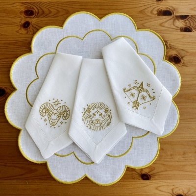 Três guardanapos brancos bordados com signos do zodíaco sobre passadeira branca com borda amarela numa mesa de madeira.