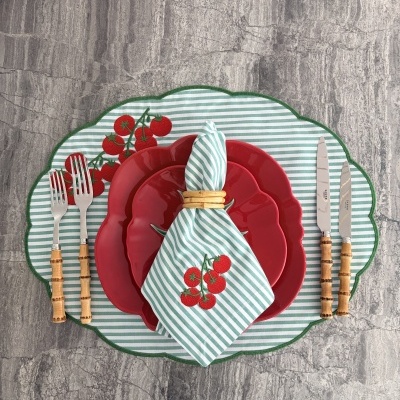 Mesa posta com pratos vermelhos, guardanapo listrado verde e branco, talheres com cabo de madeira e sousplat bordado com tomate vermelho.