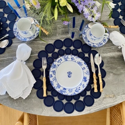 Mesa decorada para refeição com pratos brancos e azul, talheres com cabos de bambu, guardanapos brancos e arranjo floral colorido.