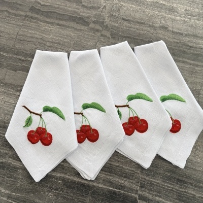 Guardanapos brancos bordados com cerejas vermelhas e folhas verdes sobre superfície cinza.