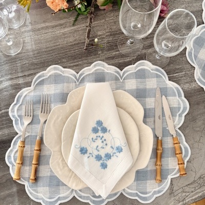 Conjunto de jantar com pratos brancos, guardanapo bordado azul, talheres com cabo de bambu, copos de vidro e base de tecido azul xadrez