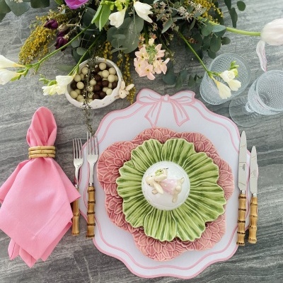 Mesa com pratos decorativos, talheres com cabo imitação bambu, guardanapo rosa, copos e flores