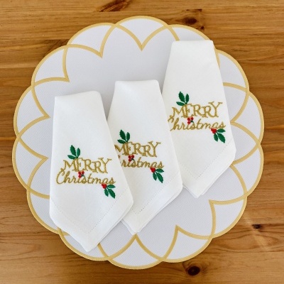 Três guardanapos brancos bordados com texto de Natal sobre prato branco com detalhes dourados