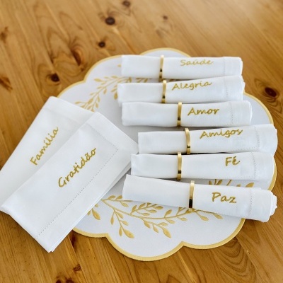 Conjunto de guardanapos brancos com palavras bordadas em dourado e suporte decorativo sobre mesa de madeira