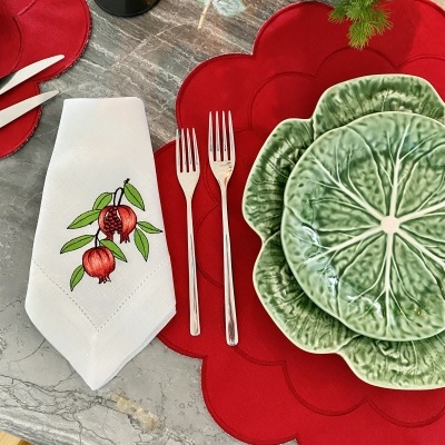 Mesa posta com pratos verdes e vermelhos, guardanapo branco bordado com romãs e talheres de metal