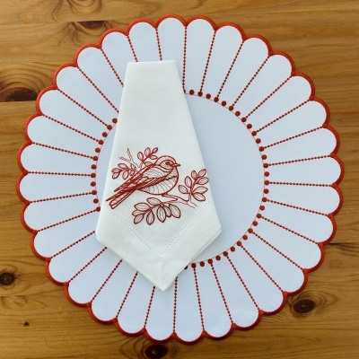 Individual redondo branco com borda vermelha e guardanapo branco bordado com pássaro vermelho sobre mesa de madeira