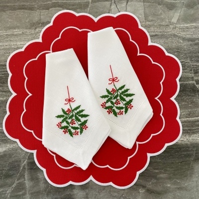 Dois guardanapos brancos bordados com azevinho sobre sousplat vermelho com pétalas brancas numa superfície cinzenta.