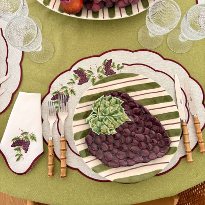 Mesa com toalha verde, prato de cerâmica decorado com cachos de uvas, talheres com cabo de madeira e copos de vidro texturizados