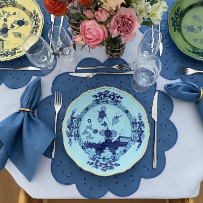 Mesa posta com prato azul claro e azul escuro, individual azul, guardanapos azuis com argolas douradas, talheres e copos de vidro, flores coloridas no centro