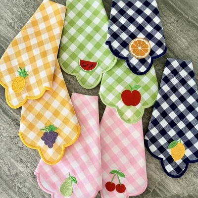 Toalhas de tecido xadrez com bordados de frutas alinhadas numa superfície de pedra cinzenta.
