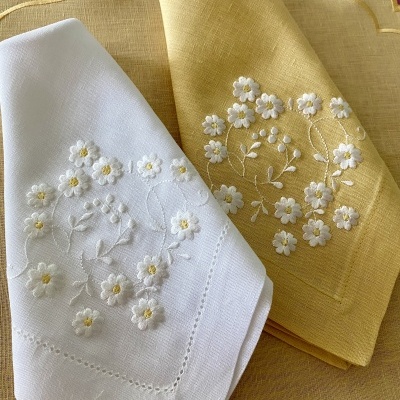 Dois panos de chá bordados, um branco e outro amarelo, com motivos florais, sobre pano amarelo com detalhes dourados